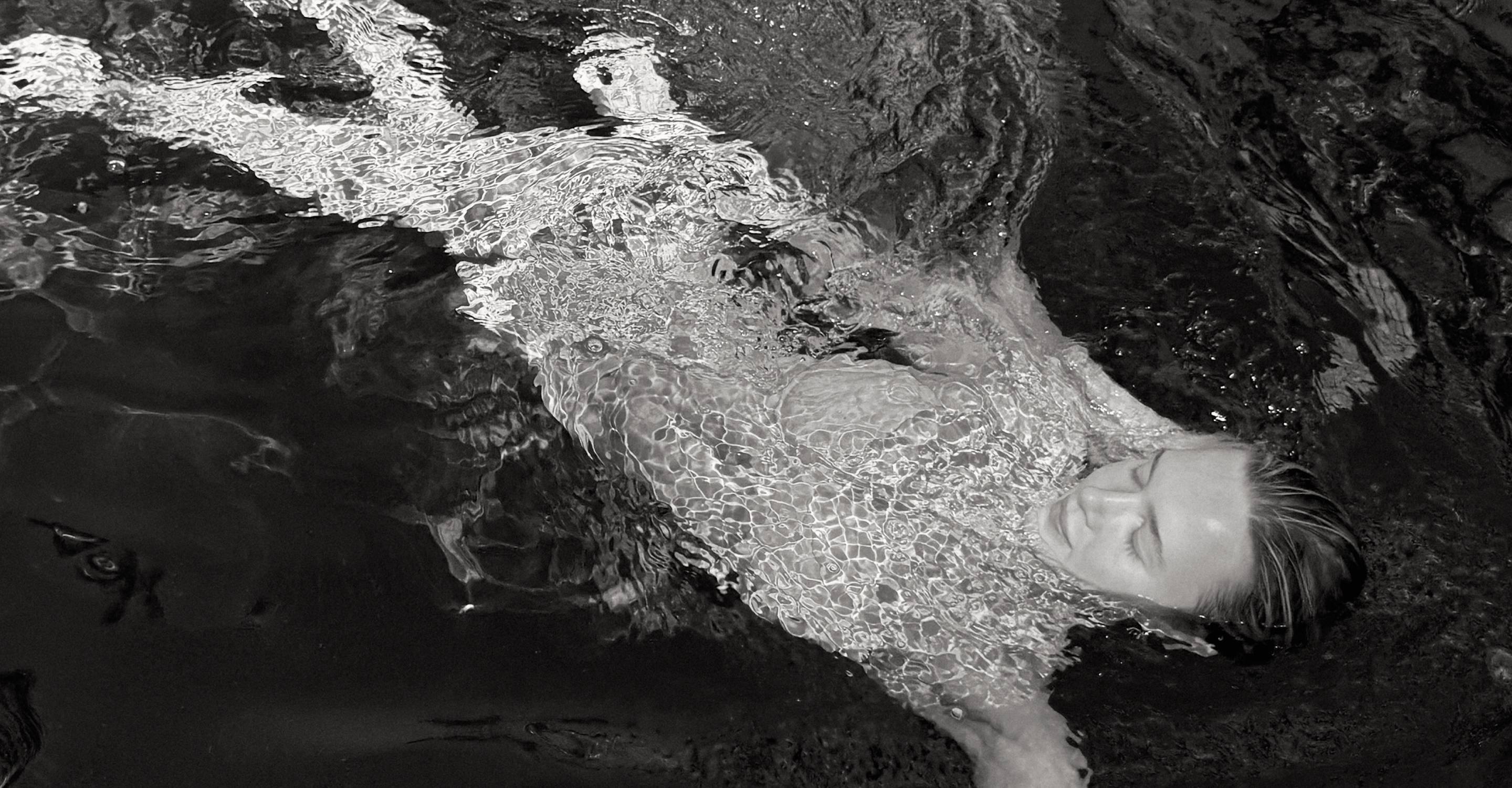 A model submerged in a pool, swimming through the surface with her face towards the camera. The woman is wearing a Wealthywet swimwear set. A black and white photo.