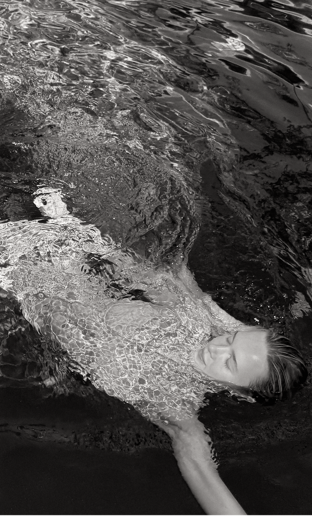 A model submerged in a pool, swimming through the surface with her face towards the camera. The woman is wearing a Wealthywet swimwear set. A black and white photo.