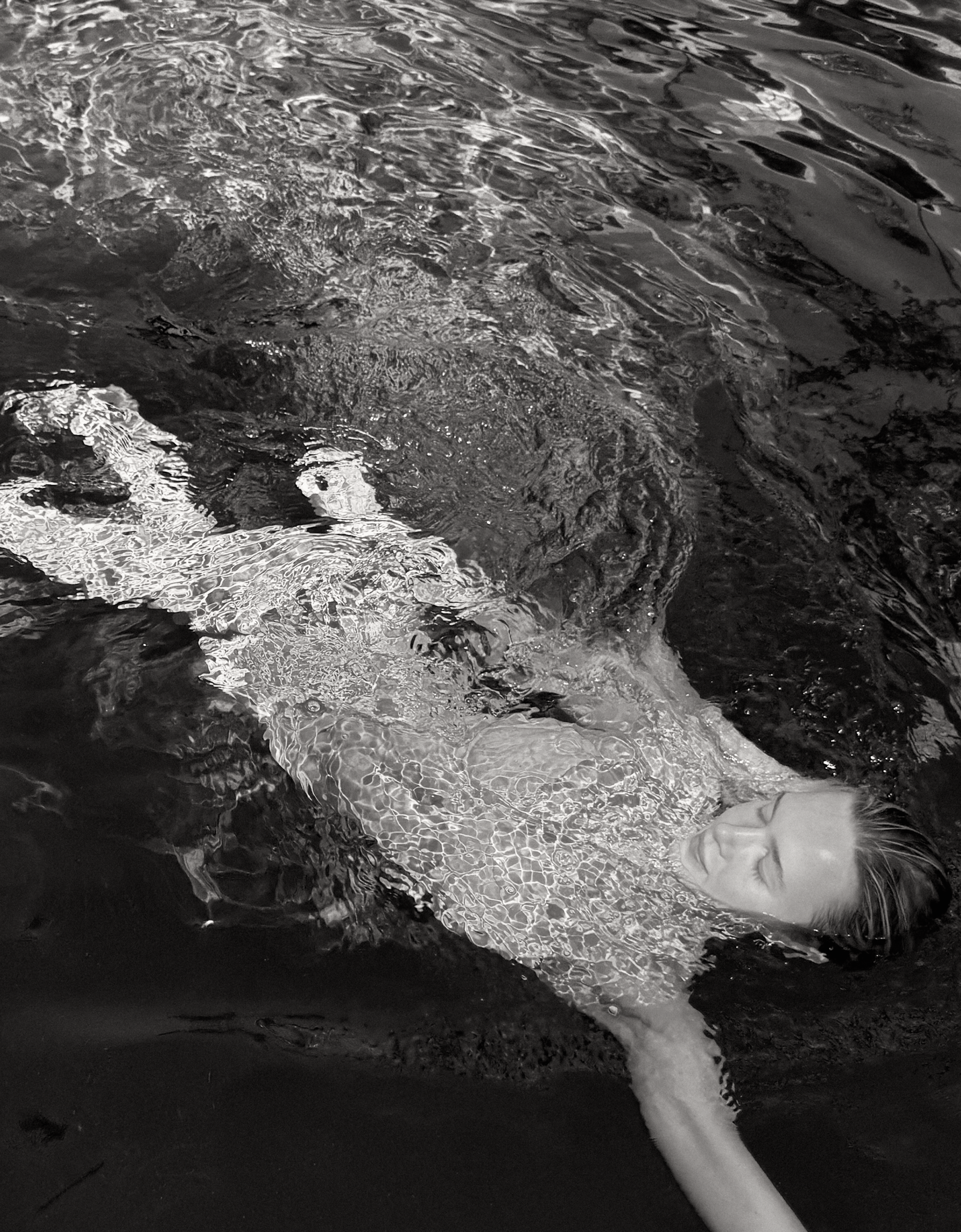 A model submerged in a pool, swimming through the surface with her face towards the camera. The woman is wearing a Wealthywet swimwear set. A black and white photo.