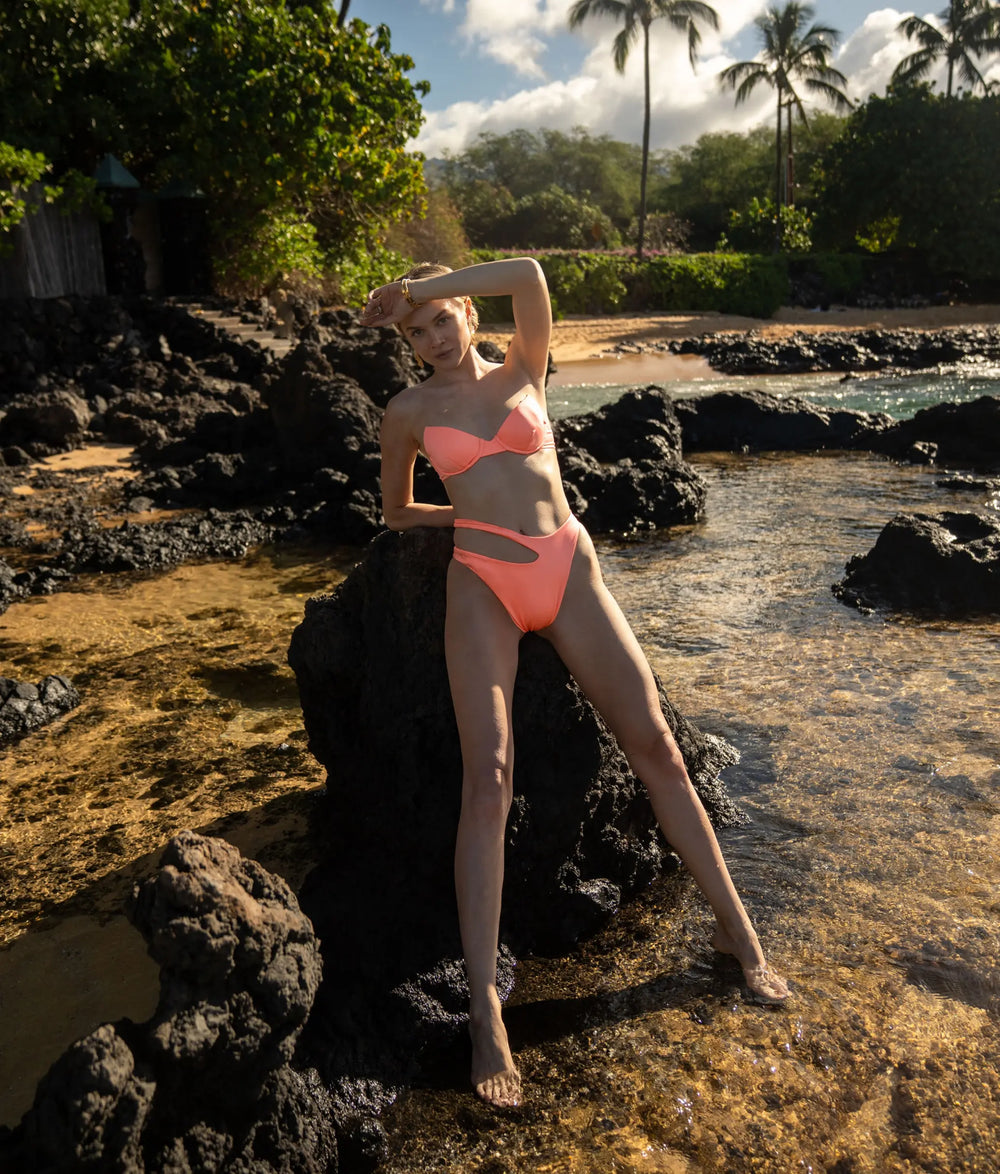 A woman is leaning  on the rock at the beach with her hand up by her head fand the palm trees in the background.  She is wearing a bright flamenco colored two piece swimwear set. The bikini top is a bandeau  structured underwire style. The bikini bottoms have an asymmetric cut out design.