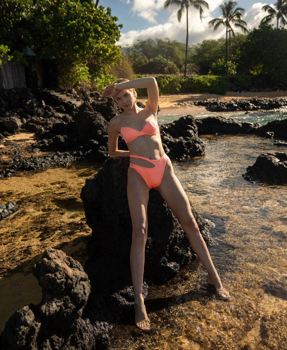 A woman is leaning  on the rock at the beach with her hand up by her head fand the palm trees in the background.  She is wearing a bright flamenco colored two piece swimwear set. The bikini top is a bandeau  structured underwire style. The bikini bottoms have an asymmetric cut out design.